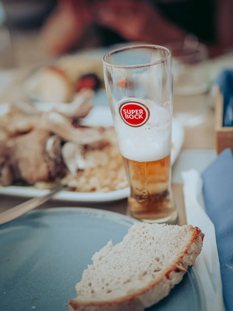 Slice of bread on a blue plate, with a partially filled glass of Super Bock beer and a meat dish in the background on a restaurant table.