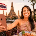 Caroline smiling taking a selfie in front of Wat Arun in Bangkok, with a Thai flag in the background.