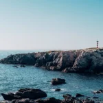 Rocky coast of Porto Covo in Alentejo, Portugal, with a red and white striped lighthouse on the cliff overlooking the Atlantic Ocean under a clear sky.