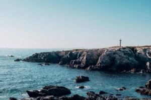 Rocky coast of Porto Covo in Alentejo, Portugal, with a red and white striped lighthouse on the cliff overlooking the Atlantic Ocean under a clear sky.