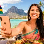 Caroline lächelt mit ihrem Smartphone vor dem Le Morne Brabant auf Mauritius, mauritische Flagge im Wind.