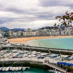 Aerial view of La Concha Bay in San Sebastián: the marina and its boats in the foreground, the shell-shaped beach, the Ferris wheel and the city in the background, autumn plane tree branch and sun rays piercing a cloudy sky, November 2021.