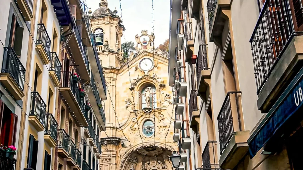 Bell tower and baroque facade of the Basilica of Saint Mary of the Choir (Santa María del Coro) in San Sebastián, viewed from below from a narrow alley in the old town lined with wrought iron balconies and fairy lights, November 2021.