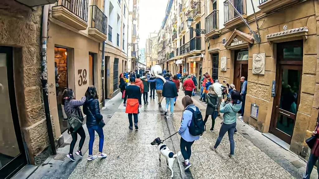 Paved and lively alley of the Parte Vieja (old town) of San Sebastián under the rain: wet cobblestones, passersby, and a waiter in a red apron between the stone buildings, November 2021.