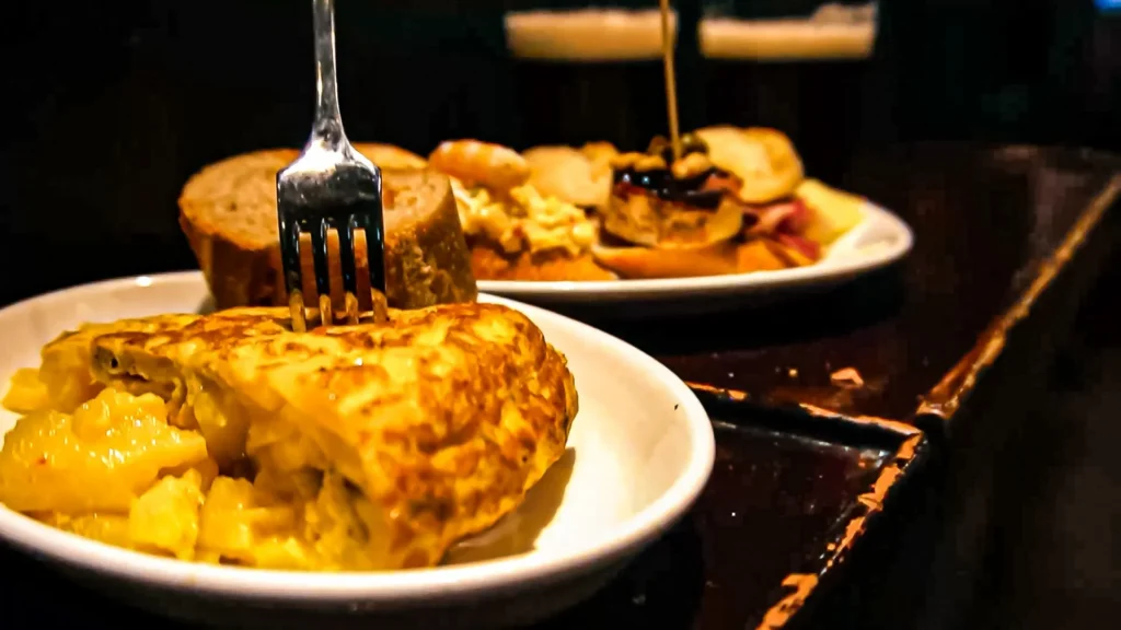 Slice of tortilla de patatas (Spanish omelette with potatoes) with a runny center, served with bread and the fork stuck in, on the dark counter of a pintxos bar in San Sebastián, November 2021.