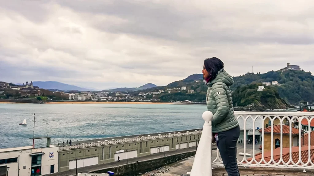 Caroline, in a teal puffer jacket, gazes at the bay of La Concha in San Sebastián from a terrace with an ornate white railing, the beach and lush green hills in the background under a gray sky, November 2021.