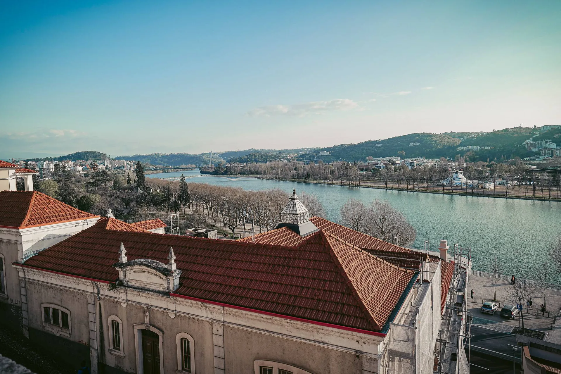 View of the red roofs of a historic building in Coimbra with the Mondego River and the surrounding hills under a clear sky.