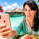 Caroline macht ein Selfie mit ihrem Smartphone vor einer Überwasser-Villa und türkisfarbenen Lagune auf den Malediven