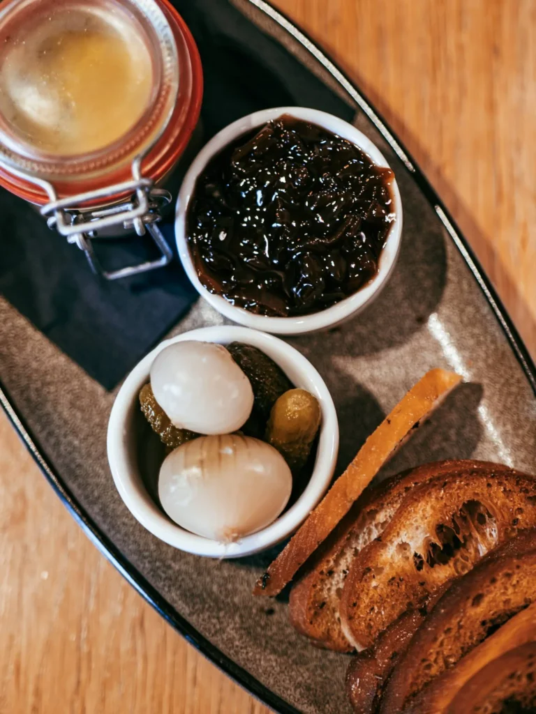 A plate composed of a jar of terrine, pickled onions and gherkins, onion jam, and slices of toasted bread, served on a wooden table at the Allan Park Hotel restaurant in Stirling in August 2026.