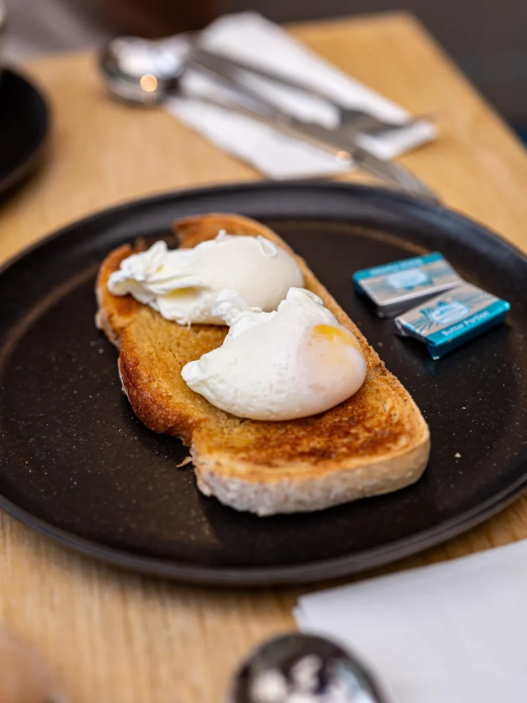 Two poached eggs served on a slice of toasted bread, accompanied by individual portions of butter on a black plate, on a wooden table in a room at the Allan Park Hotel in Stirling in August 2026.