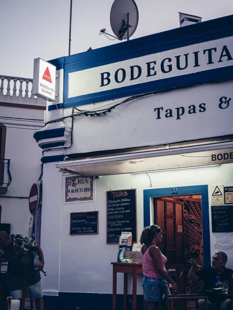 Facade of Bodeguita Salinas in Tavira, Algarve, tapas and wine bar on Rua 4 de Outubro: Caroline standing on the terrace and Xavier seated at the table, summer evening atmosphere.