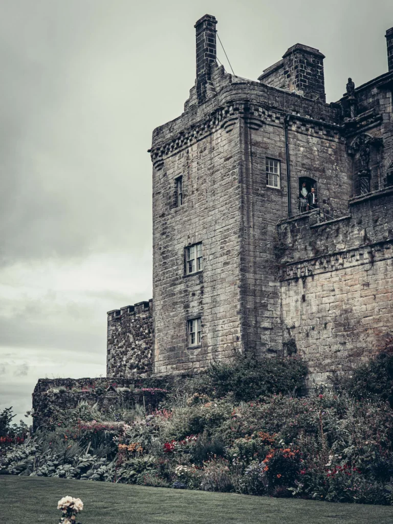 The stone front of Stirling Castle, Scotland, with a flower garden in the foreground under a cloudy sky.