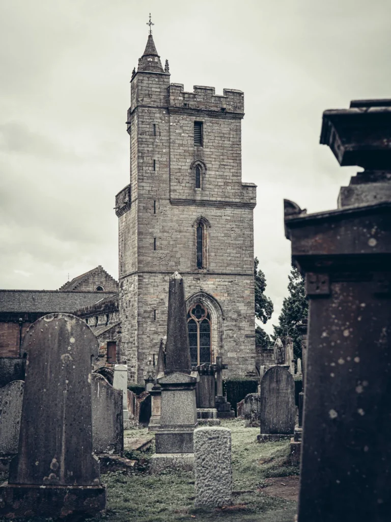 Exterior of the Church of the Holy Rude, in Stirling, with its stone steeple and the kirkyard where grass grows around the headstones. The church is located on St John Street, just below the castle.