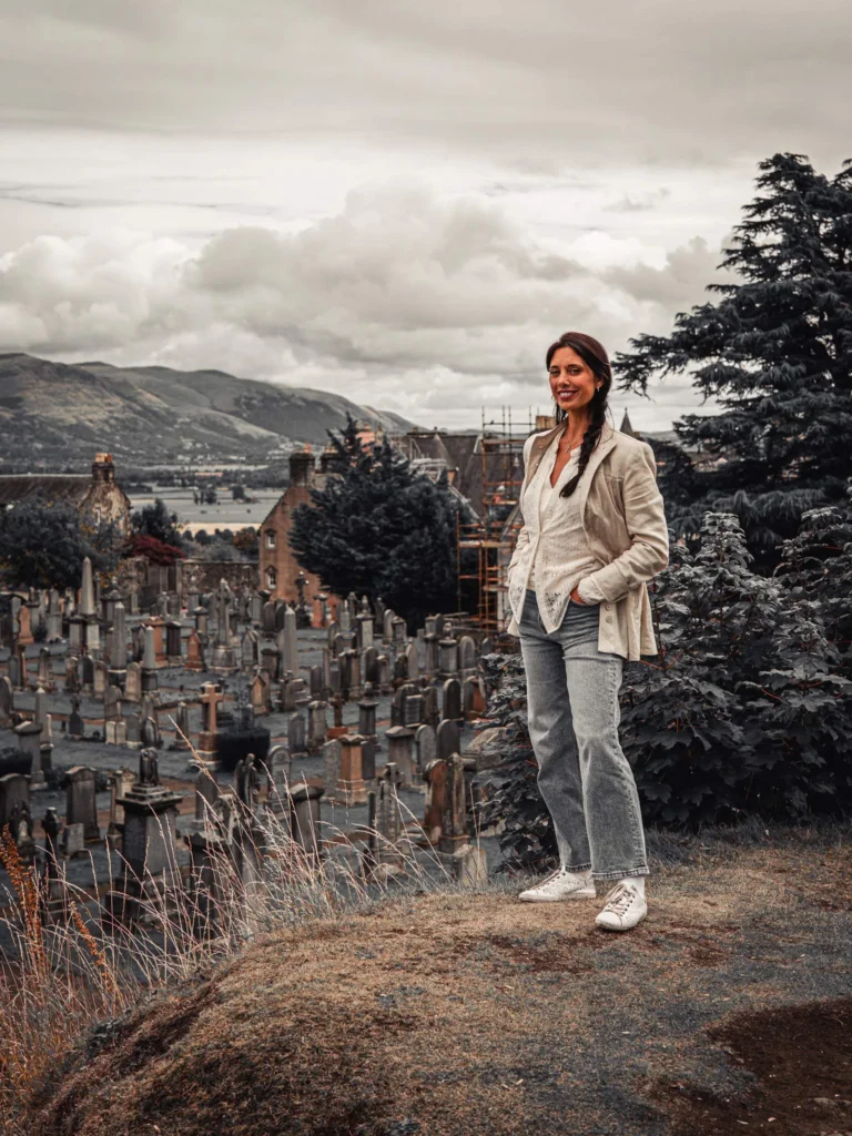 Caroline on the hill of the kirkyard of the Church of the Holy Rude, in Stirling, with a view of the countryside and the grass-covered gravestones.