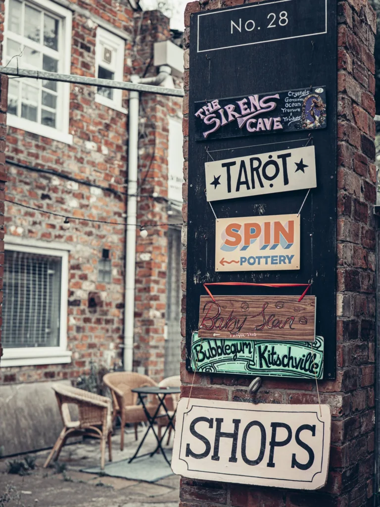 Signage panel of unit No. 28 of The Hidden Lane, in Glasgow: The Sirens Cave (crystals, gemstones, ocean treasure & trinkets), Tarot, Spin Pottery, Bubblegum Kitschville, and a vintage shop, under a "SHOPS" sign. Behind, the small terrace with wicker chairs.
