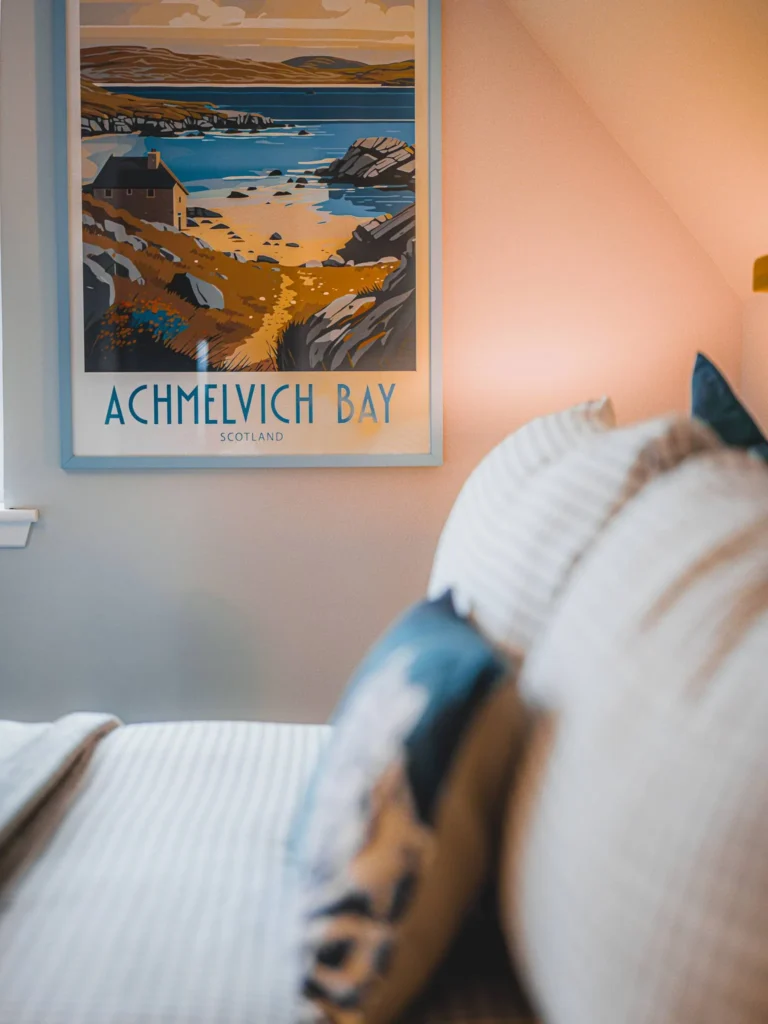 Colourful poster of Achmelvich Bay hanging on the wall of a cosy room, with a bed and cushions in the foreground. Warm atmosphere in a Scottish Highlands accommodation.