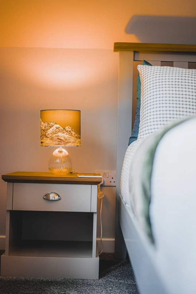 Light wood bedside table with lamp on and tartan patterned bed in a room in Glencoe, Highlands, Scotland.
