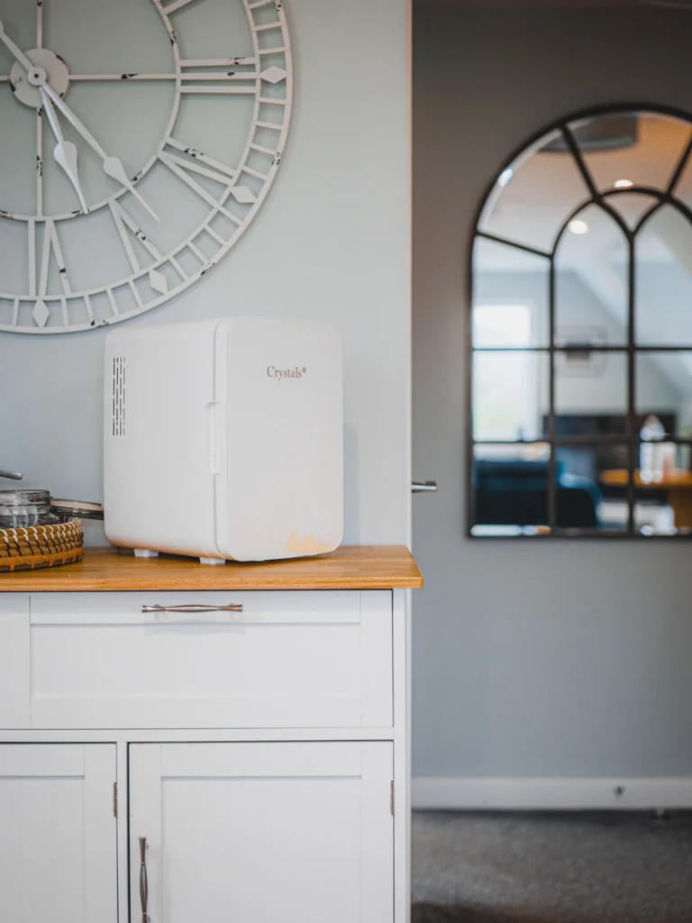 Small white fridge from the brand Crystals placed on a light wood cabinet in a bright room, with a large wall clock and a black-framed mirror reflecting the interior of the accommodation in Glencoe in the Scottish Highlands.