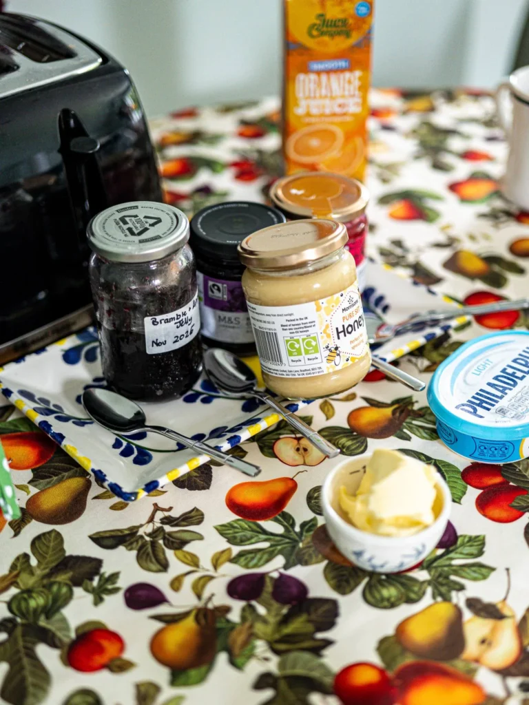 Scottish breakfast with jam, honey, butter, cream cheese, and orange juice on a fruit-patterned tablecloth, probably at Rocklee Bnb in the Highlands in August 2026.