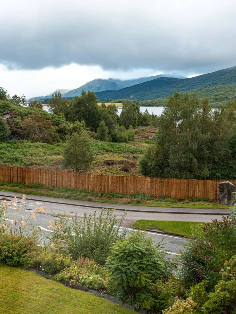 View of a lush garden with a road, a wooden fence, and in the background, a landscape of mountains and lochs typical of the Scottish Highlands under a cloudy sky.