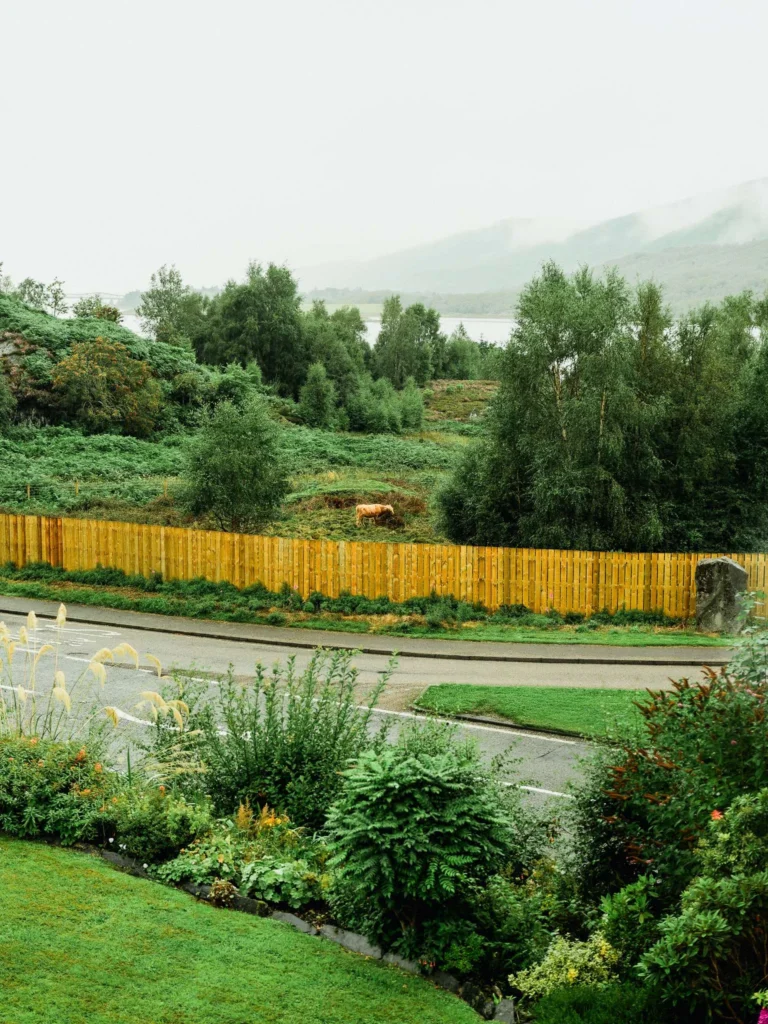 View of a lush garden with a wooden fence, wooded hills, and a lake in the background under a misty sky in the Scottish Highlands.