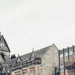 Paved sloping street of the old town of Stirling, lined with large stone and sandstone buildings, a monument and a clock in the distance. The town seems less r