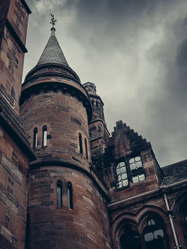 Stone facade of the University of Glasgow, tower and mullioned windows under a cloudy sky.