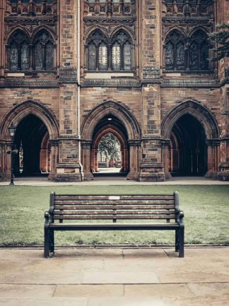 Wooden bench in front of an archway and stained glass facade of the University of Glasgow, in the green courtyard served by the Cloisters passage.
