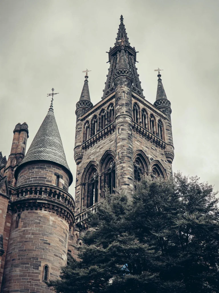 Tour of the Main Building of the University of Glasgow and its soaring spires, partially obscured by the trees under a cloudy sky.