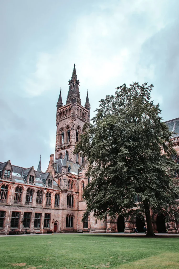 View of the inner courtyard and the Gothic tower of the University of Glasgow, with a large lawn and a tree in the foreground, under a cloudy sky.