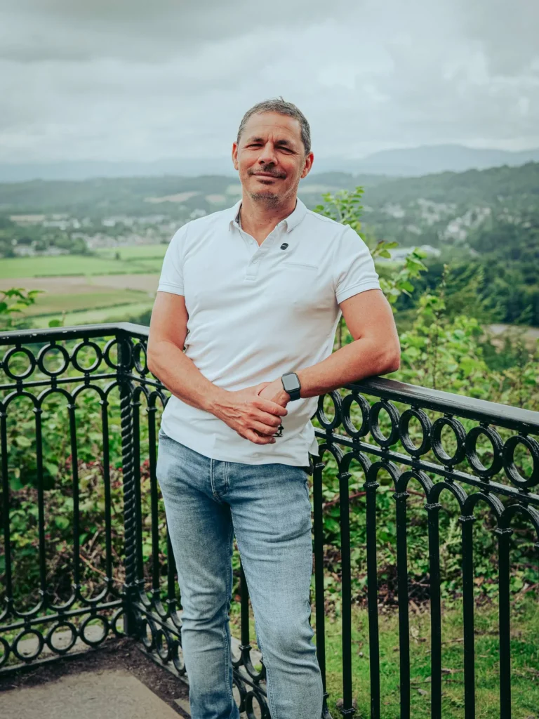Xavier poses in front of a wrought iron railing with a panoramic view of the Scottish countryside near the Wallace Monument in Stirling, under a cloudy sky in August 2026.
