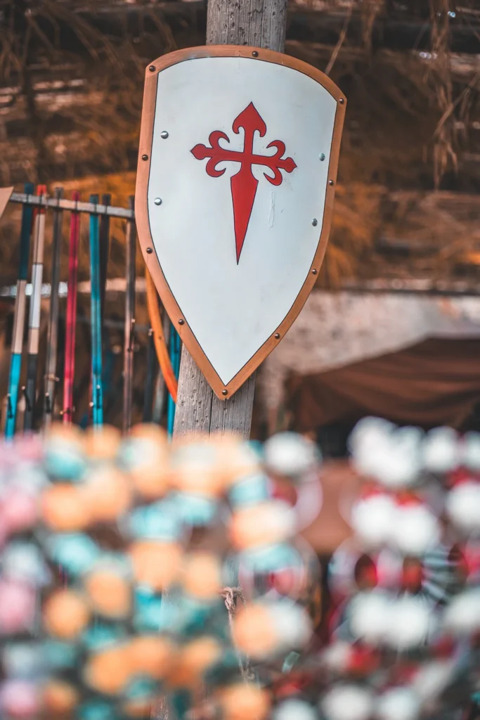 A medieval shield decorated with a red cross is hanging on a wooden post, with colorful accessories in the background during a historical festival.