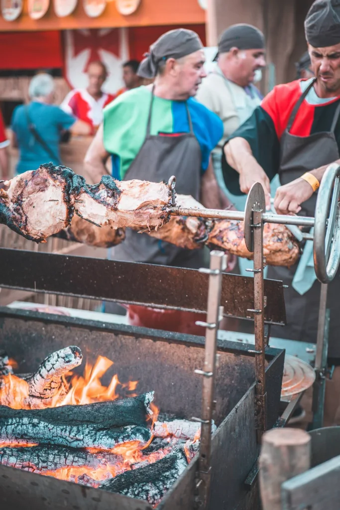 Men in medieval costumes are roasting large cuts of meat on a spit over a wood fire during a street performance, in a festive historical atmosphere.